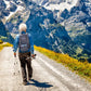 Gray Inogen One G5 backpack with logo worn by a hiker on a mountain trail surrounded by wildflowers and snowy peaks.